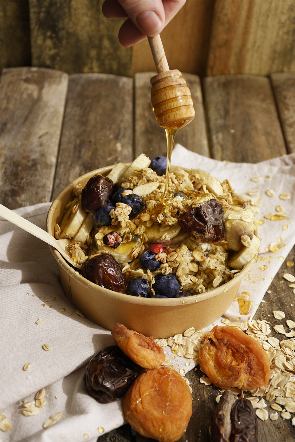 A bowl of overnight oats with mixed fresh and dried fruits sitting on a neutral coloured napkin on a wooden table and a hand holding a wooden honey dipper drizzling honey over the bowl